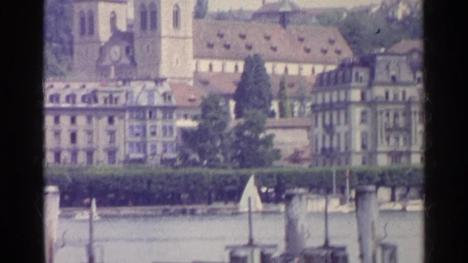 1961 8mm view of Swiss lakeside buildings with a twin-spired church, tiled rooftops, trees, and waterfront structures.