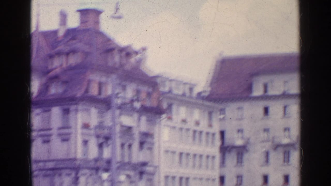Faded 1961 8mm frame of Swiss town buildings with ornate rooflines, dormers, pale facades, and overhead street wires.