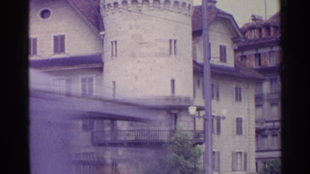 1961 Switzerland 8mm frame showing the lower portion of a stone tower, pale building facades, balconies, streetlamps, and a pole.