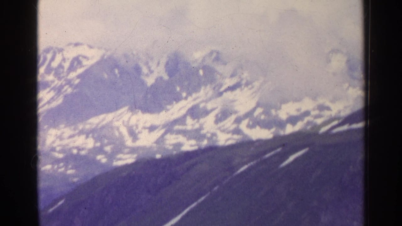1961 Switzerland 8mm frame showing distant snowy alpine peaks beneath an overcast sky, with soft focus and faded blue-purple color.