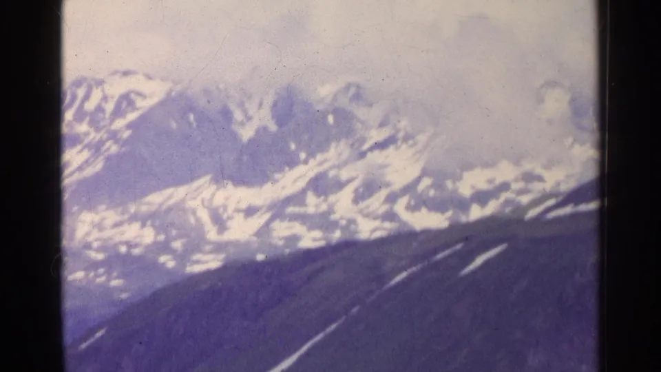 1961 Switzerland 8mm frame showing distant snowy alpine peaks beneath an overcast sky, with soft focus and faded blue-purple color.