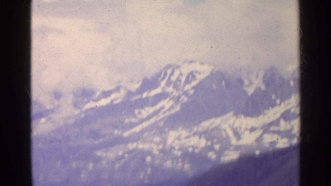1961 Switzerland home-movie frame with hazy snow-streaked alpine peaks, muted exposure, and visible 8mm film texture.
