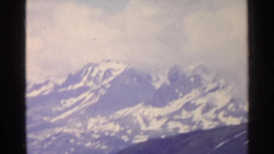 Final 1961 Switzerland frame showing snow-capped alpine mountains, dark foreground slopes, overcast sky, and aged 8mm frame markings.