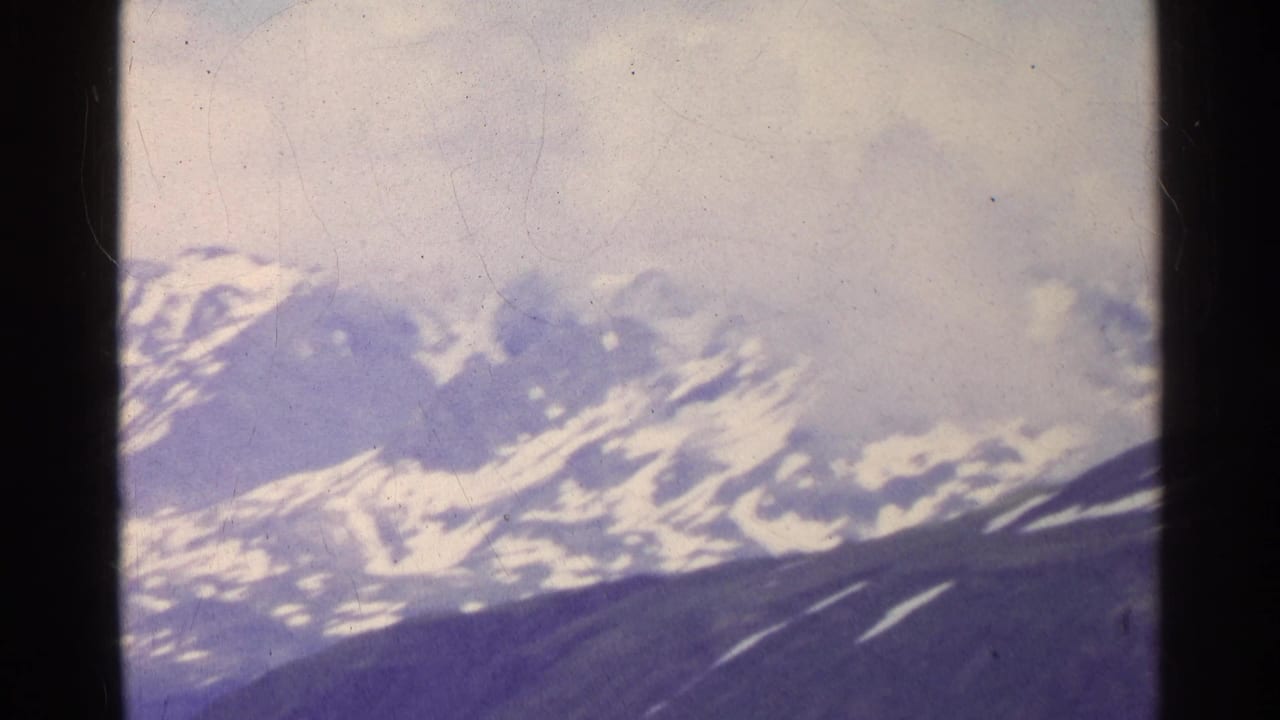 1961 Swiss 8mm view of distant snow-covered mountains under clouds, seen through soft film grain and dark frame borders.