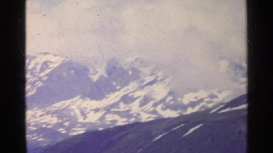 1961 Swiss 8mm view of distant snow-covered mountains under clouds, seen through soft film grain and dark frame borders.