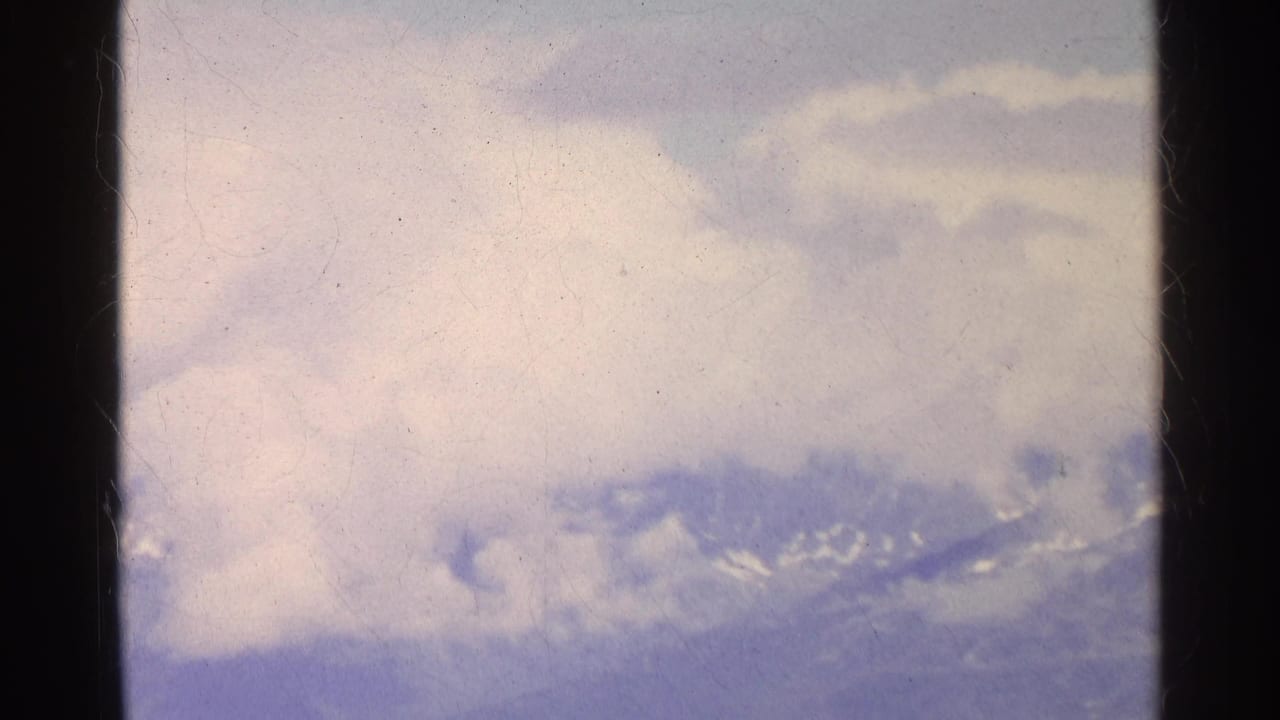 1961 Swiss alpine landscape with a hazy snow-dusted ridge and bright cloud cover in a soft 8mm home-movie frame.