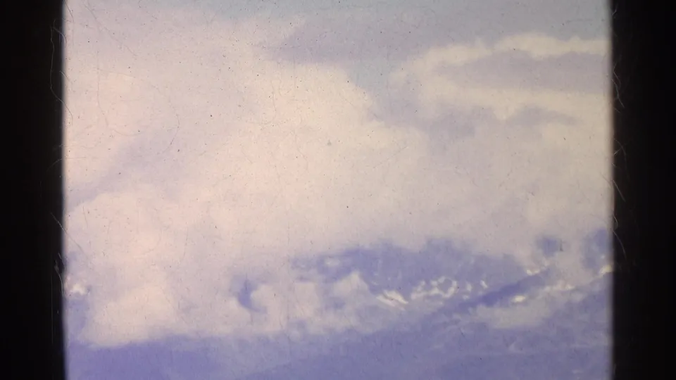1961 Swiss alpine landscape with a hazy snow-dusted ridge and bright cloud cover in a soft 8mm home-movie frame.