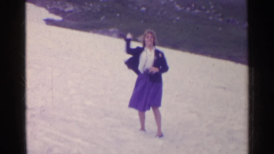 A woman in a dark jacket and purple skirt raises her arm while standing on snow in the 1961 Switzerland footage.