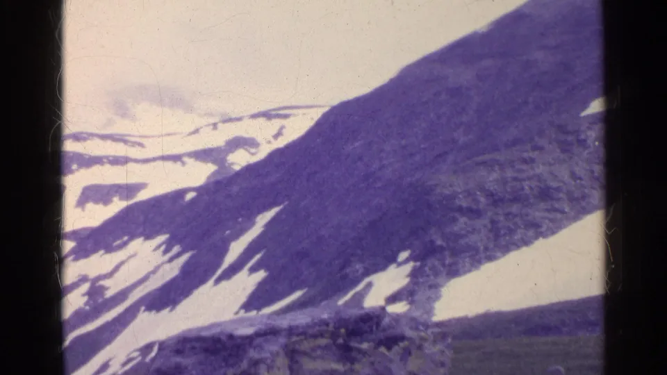 1961 Switzerland home-movie frame of an alpine slope with melting snow patches and a distant snow-covered ridge.