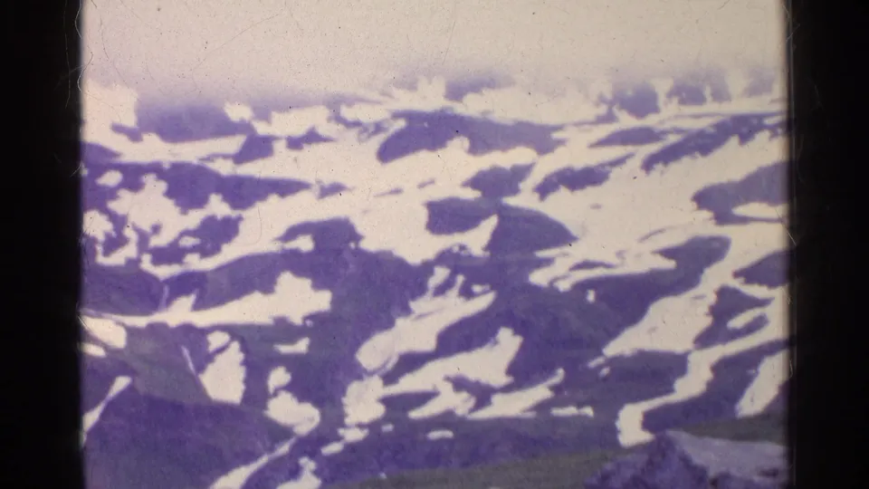 Aerial 8mm frame from Switzerland showing snow-streaked alpine ridges beneath pale haze.