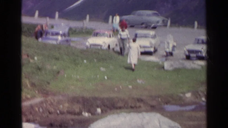 Tourists stand and walk near parked cars on a Swiss mountain road in 1961 home-movie footage.