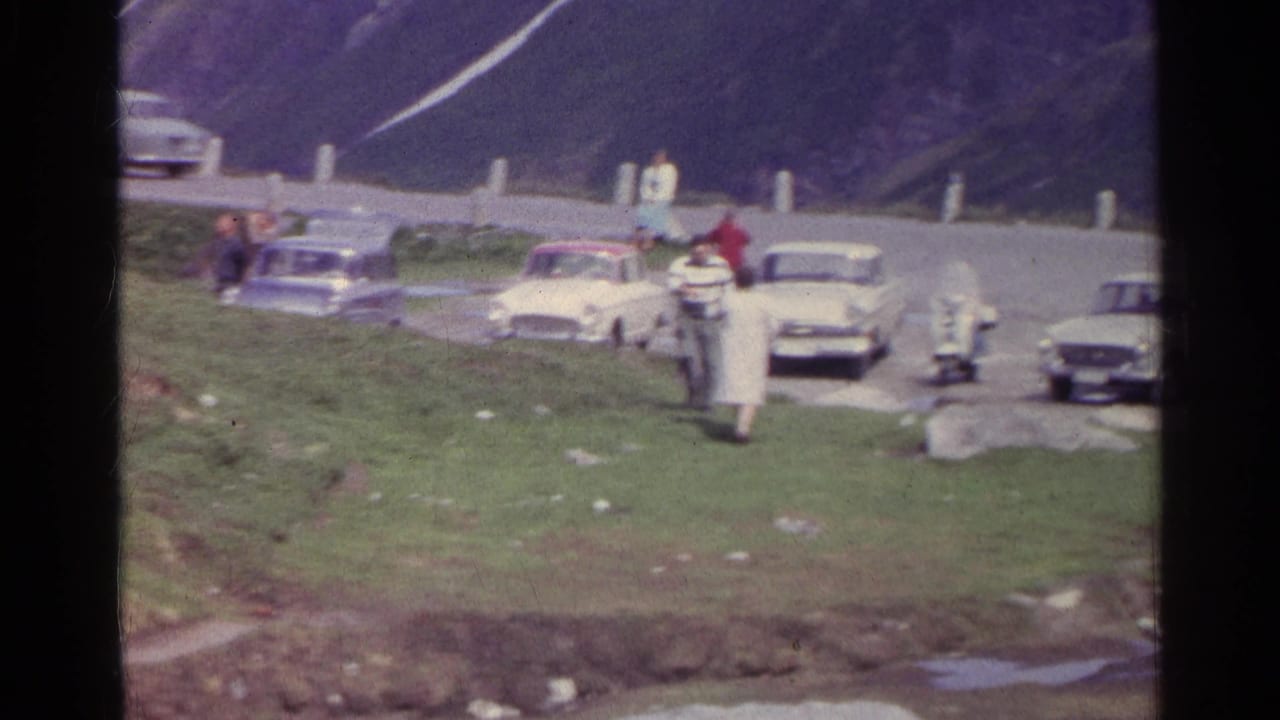 A visitor walks toward the camera while others gather near classic cars on a Swiss alpine road.