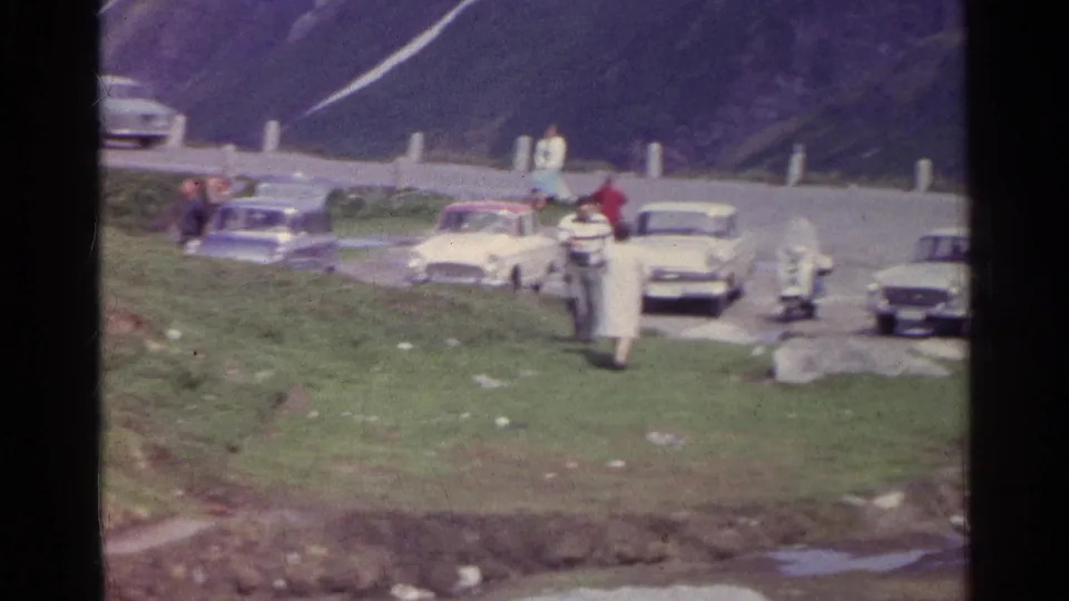 A visitor walks toward the camera while others gather near classic cars on a Swiss alpine road.