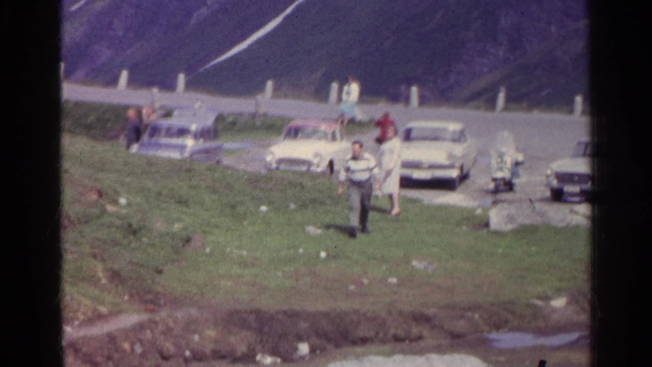 1961 Swiss mountain-road stop with tourists, roadside posts, grass, rocks, and parked cars in soft 8mm color.