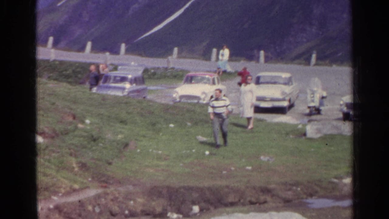 Man in a striped sweater walks down a grassy hillside near parked cars and bystanders.