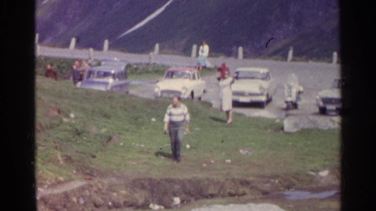 The man walks toward the camera across grass, with several people and mid-century cars visible along the Swiss mountain road.