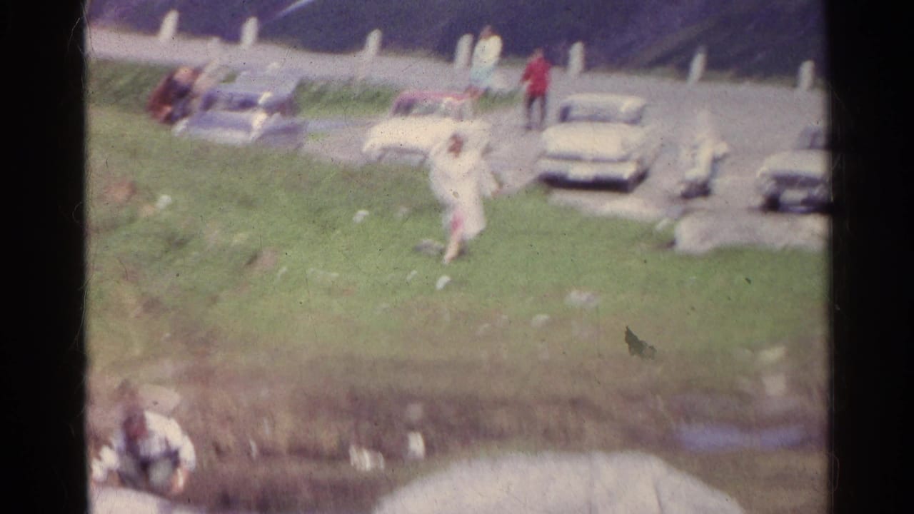 A soft, blurred 8mm frame shows the woman in white moving on the hillside near parked cars and the mountain road.