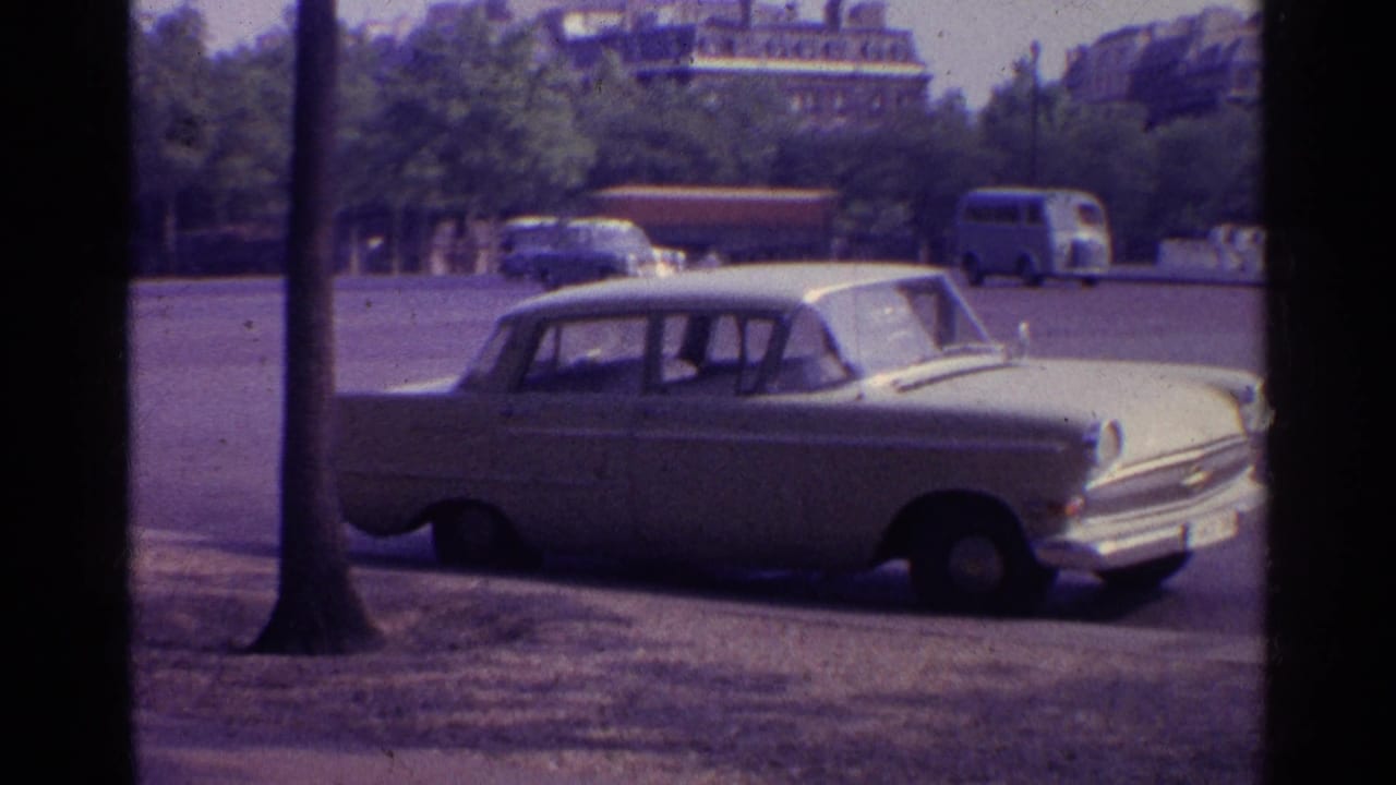 1961 8mm frame of a white sedan parked near a tree on a European street, with buses, cars, greenery, and older buildings beyond.