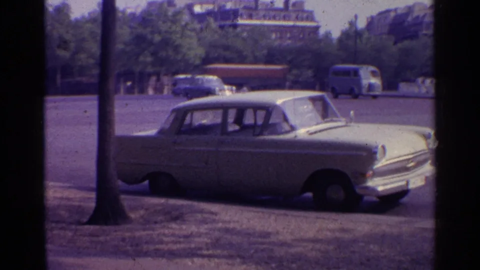 1961 8mm frame of a white sedan parked near a tree on a European street, with buses, cars, greenery, and older buildings beyond.