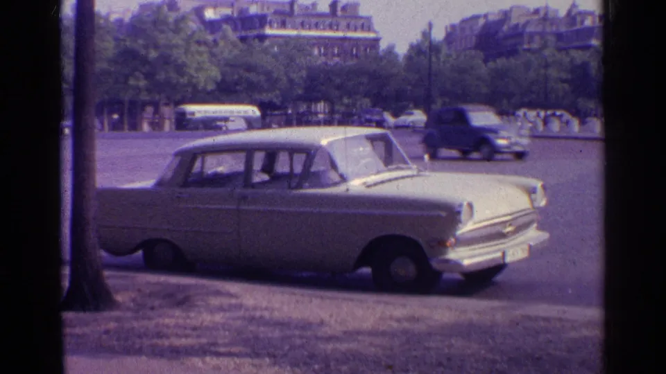 A white 1960s-era sedan remains parked near the curb while background vehicles pass across a tree-lined urban roadway.