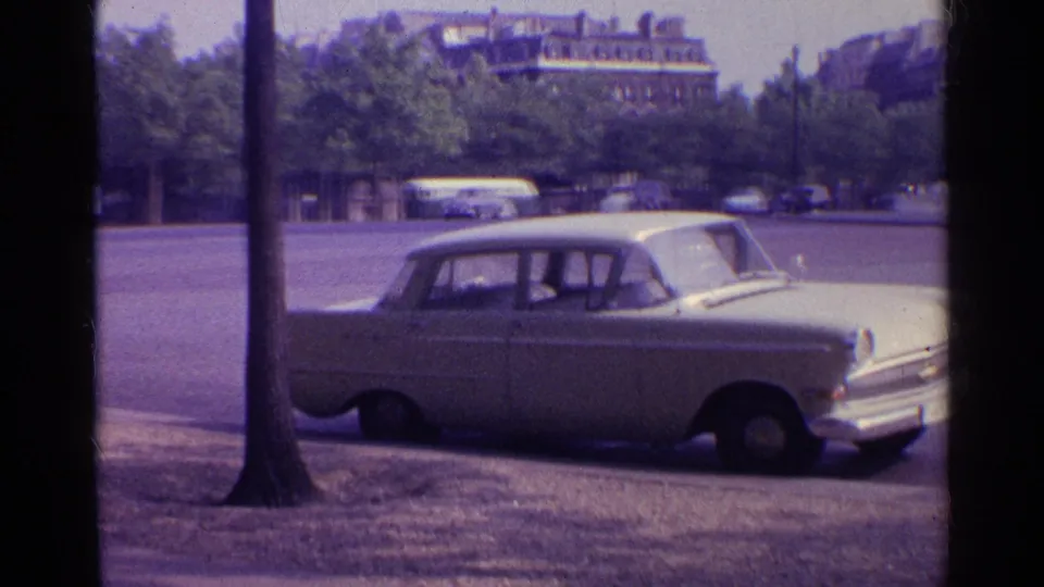 The white sedan fills the lower frame in 1961 8mm footage, with a bus, cars, trees, and urban buildings beyond the roadway.