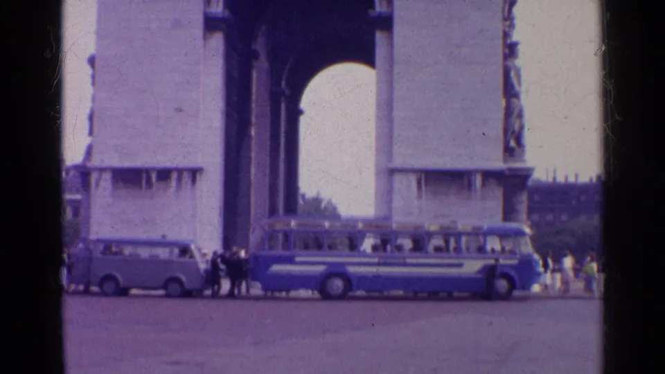 A large stone arch monument appears beyond the road, with a blue bus, small van, and distant pedestrians visible at street level.