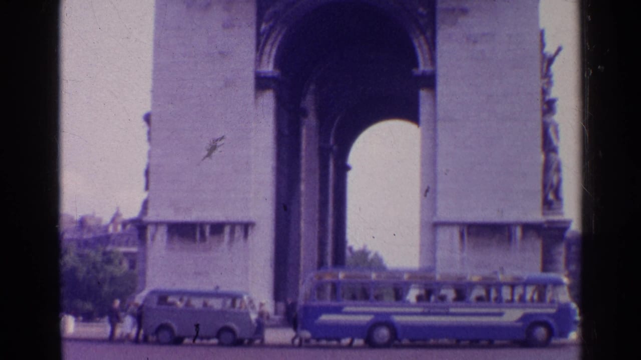 A blue coach and light van sit near the base of a tall stone arch in 1961 Switzerland, with visitors visible at a distance.