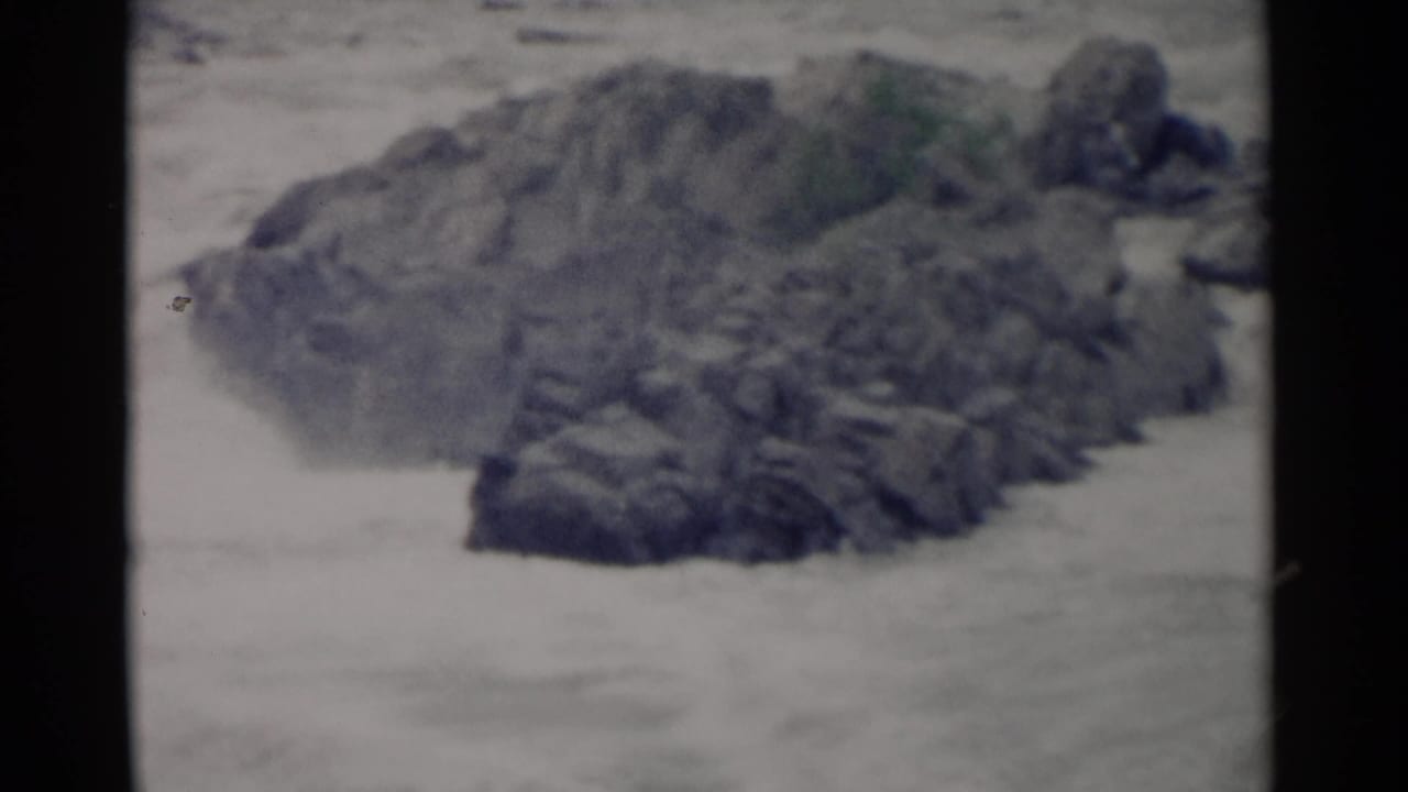1962 Jamestown, Virginia home-movie frame of whitewater moving around a dark rocky outcrop, with visible 8mm grain and gate edges.