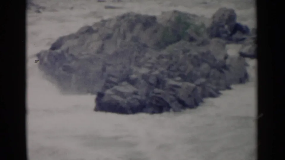 1962 Jamestown, Virginia home-movie frame of whitewater moving around a dark rocky outcrop, with visible 8mm grain and gate edges.