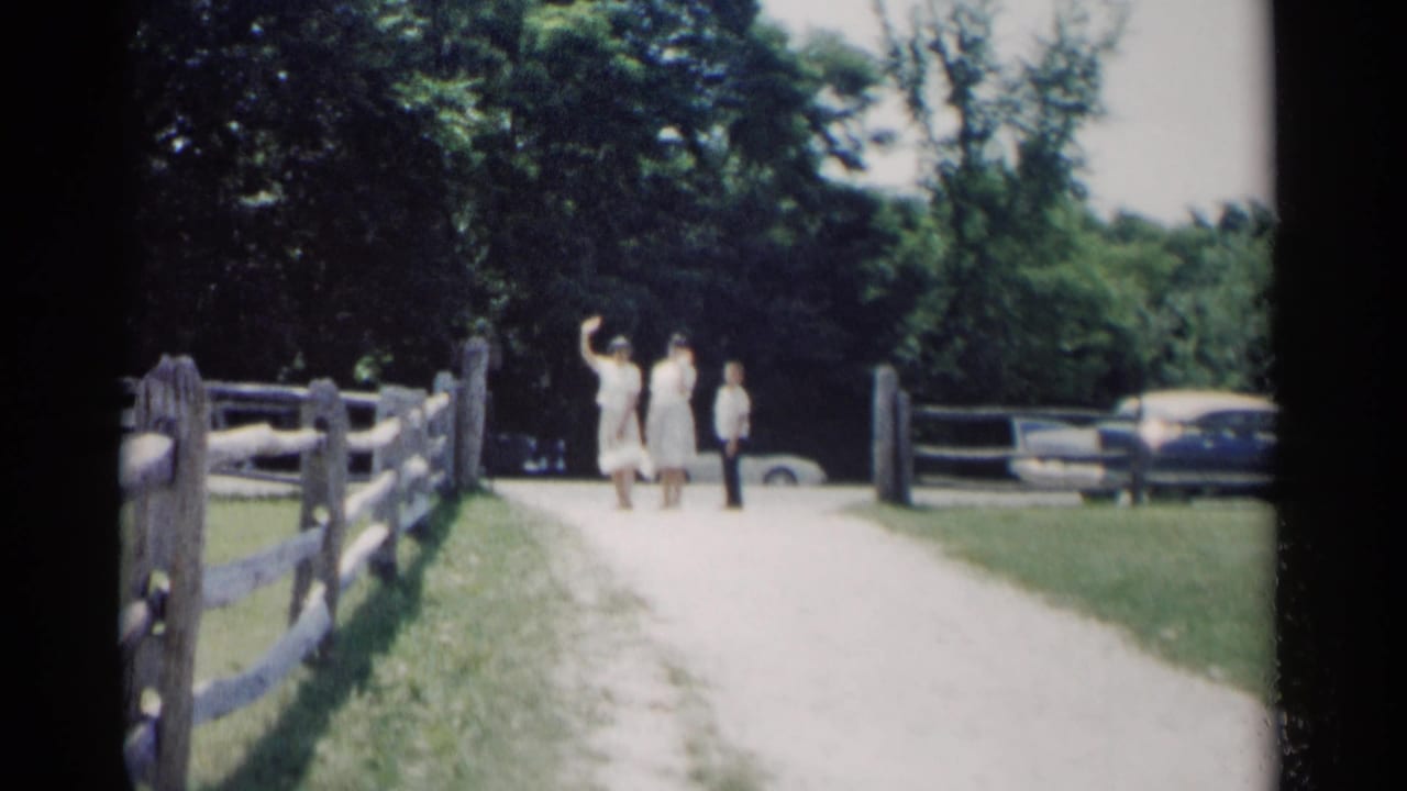 Two young women and a child-sized figure walk on a gravel path beside a split-rail fence.