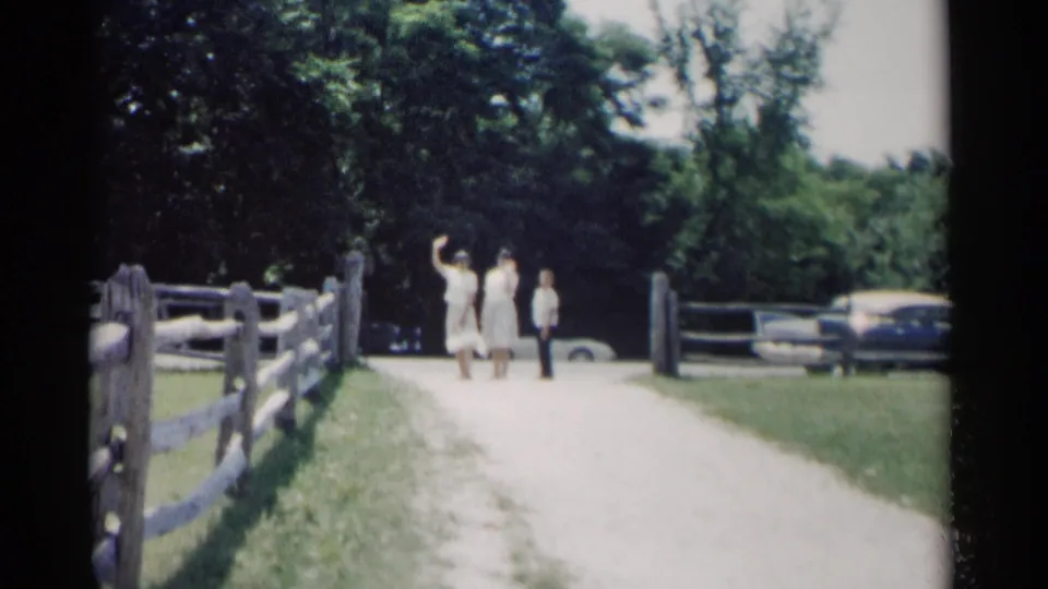 1962 Jamestown, Virginia home-movie frame of visitors on a gravel path near trees, fencing, and parked cars.