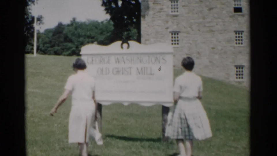 Two young women approach the George Washington's Old Grist Mill sign in Jamestown, Virginia, with a stone building behind it.