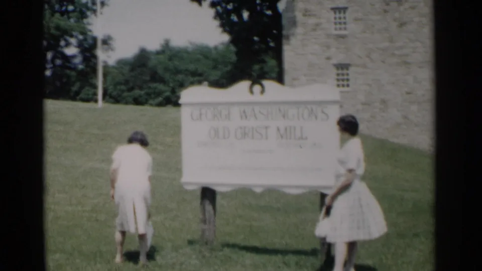 At George Washington's Old Grist Mill, one visitor bends near the lawn as another stands by the sign and stone building.