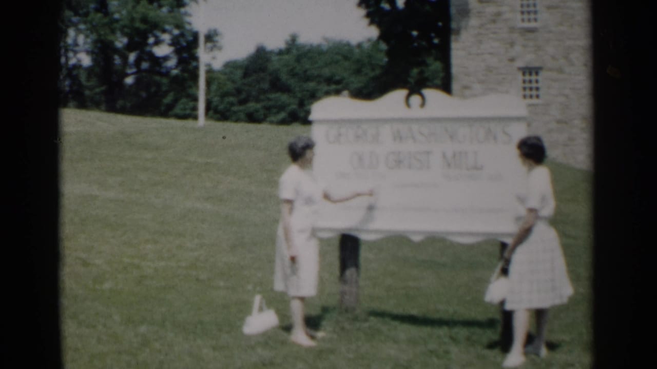 In 1962 Jamestown, Virginia footage, two women in light dresses pose beside the George Washington's Old Grist Mill sign near a stone building.