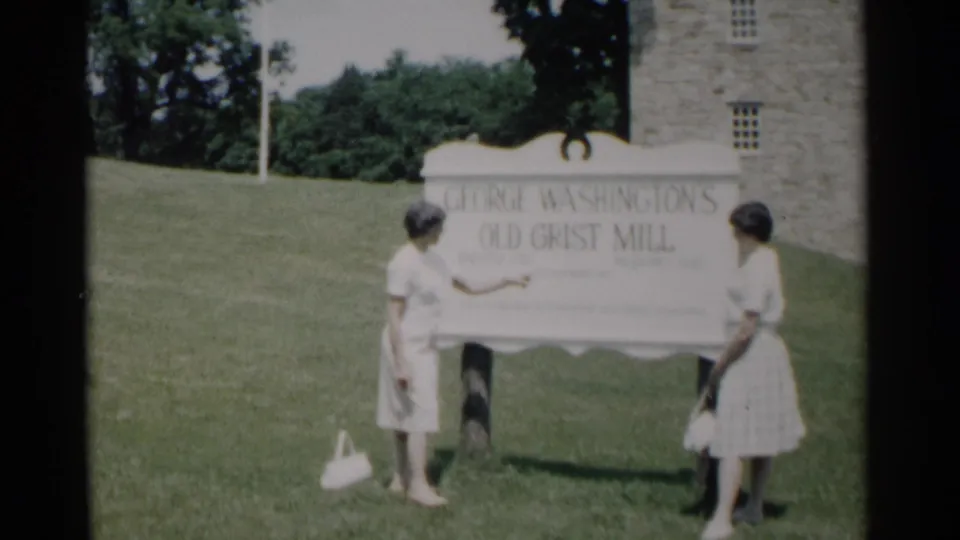 Two women in summer dresses stand in front of George Washington's Old Grist Mill sign at the Jamestown historic site.