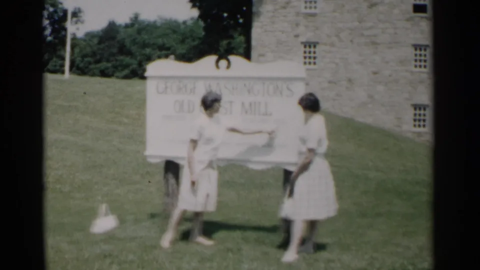 One woman gestures toward the grist mill sign while another stands beside her on the lawn in 1962 Jamestown footage.