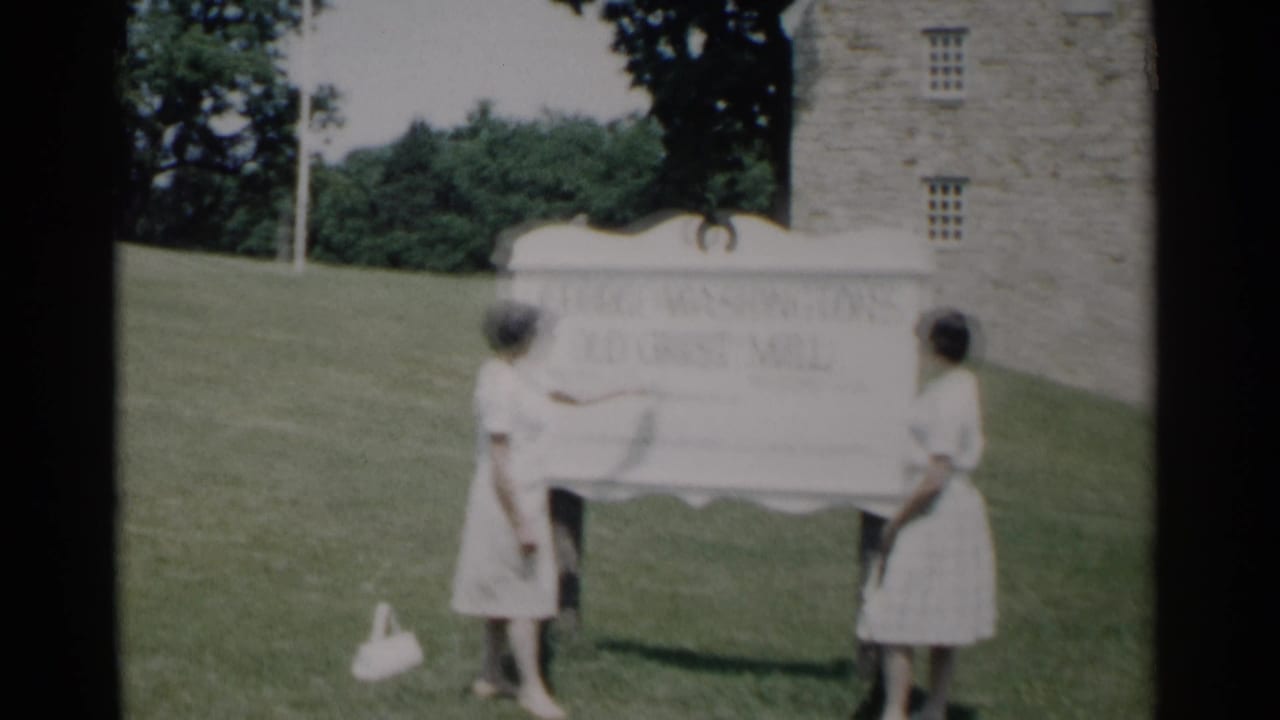 Two young women stand beside a white grist mill sign on a grassy lawn.