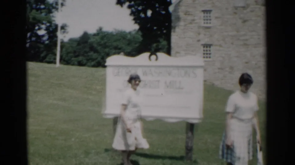 The women move past the George Washington's Old Grist Mill sign in a sunny Jamestown, Virginia scene, with the stone mill wall in the background.