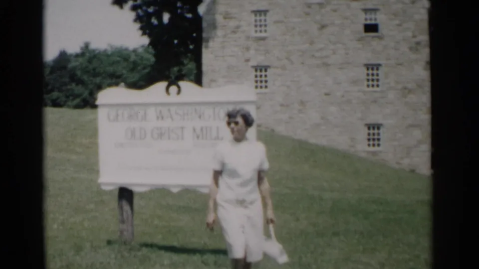 A young woman in light clothing stands before the grist mill sign on a green lawn, with the stone mill building visible behind her.