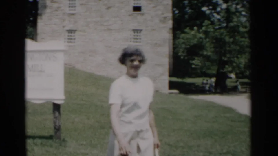 A woman in light clothing approaches the camera in the Jamestown grist mill scene, with the sign partly cropped and the stone building behind her.