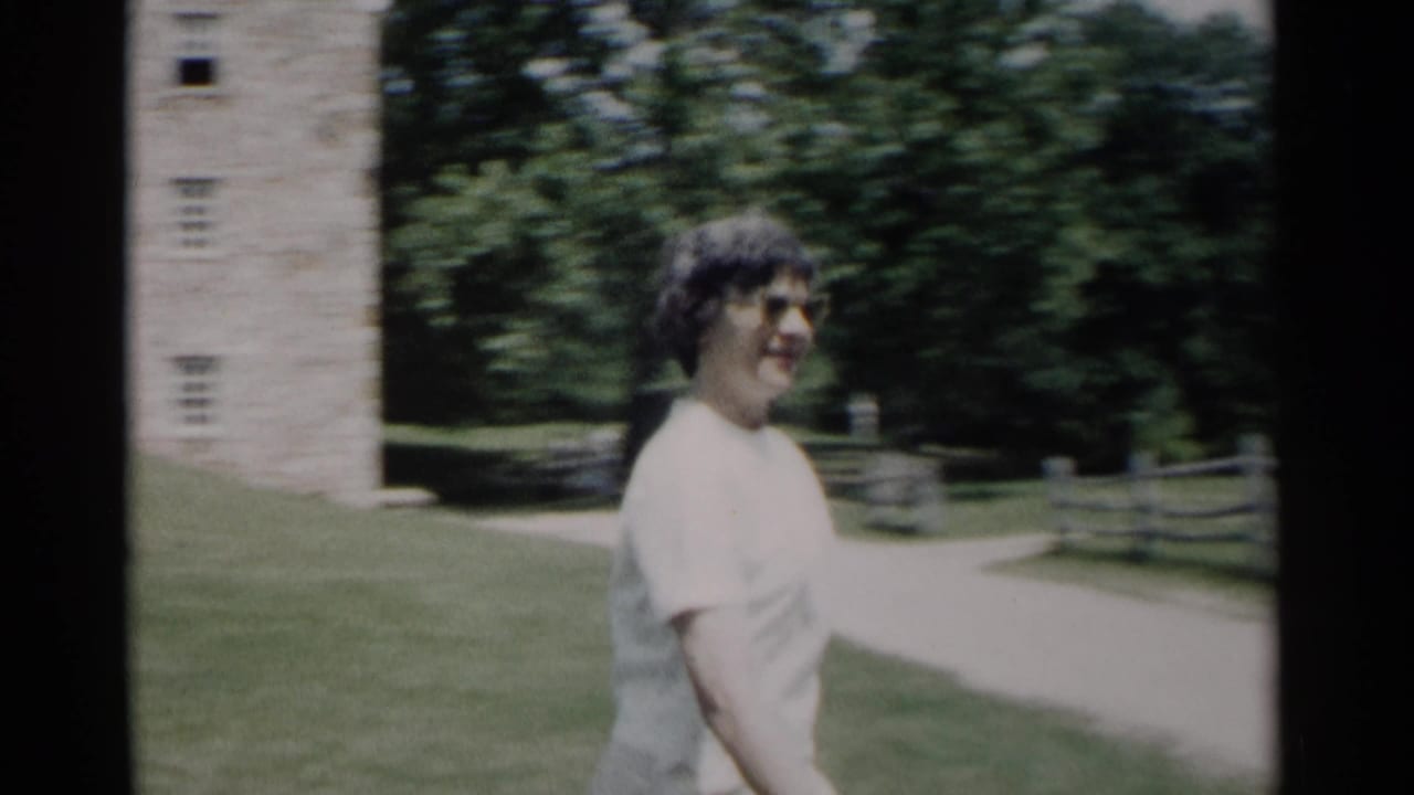 In Jamestown, Virginia, a woman in a white dress and glasses walks beside a path near a stone structure and trees in 1962 home-movie footage.