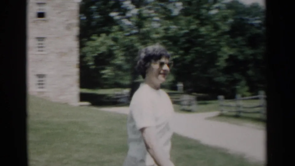 In Jamestown, Virginia, a woman in a white dress and glasses walks beside a path near a stone structure and trees in 1962 home-movie footage.