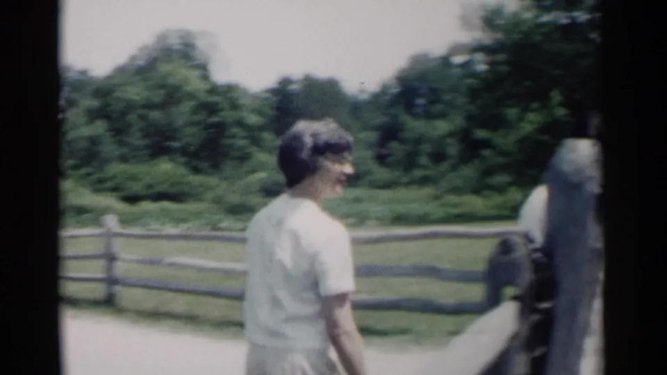 A woman in a white dress and glasses walks along a Jamestown path near a split-rail fence and gate area in 1962 8mm footage.