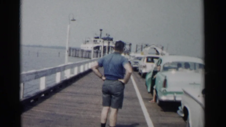 A man in shorts stands with hands on hips on the Jamestown pier, with parked cars, passengers, water, and a ferry ahead.