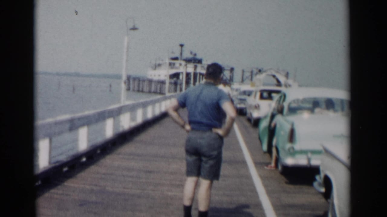 Man in blue shirt stands on a pier while cars line up near a ferryboat.