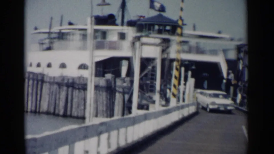 A car moves at the ferry landing while the ferry structure, pier railing, and water are visible in 1962 Jamestown footage.