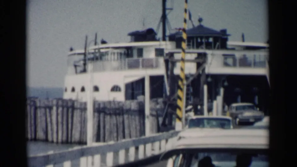 1962 Jamestown Ferry scene showing a white ferry docked at the ramp as early-1960s cars drive off toward the camera.