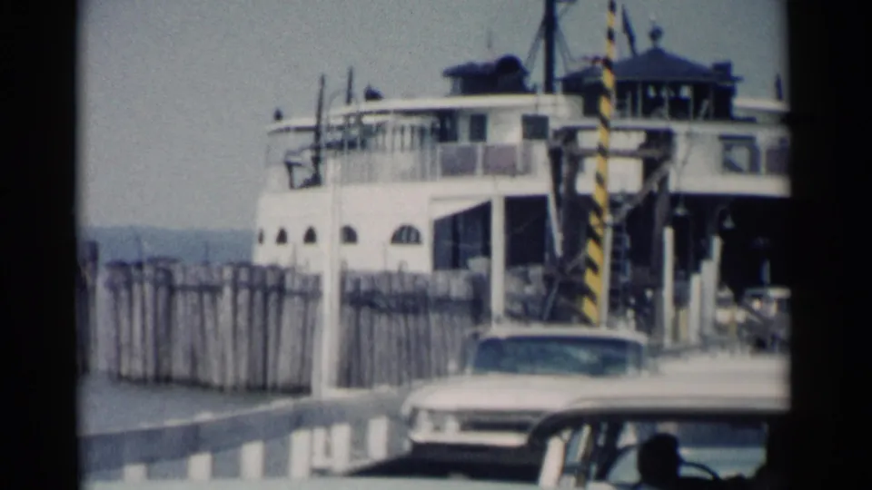 Jamestown Ferry disembarkation view with a white car approaching the camera, striped ramp posts, pier fencing, and the ferry behind.