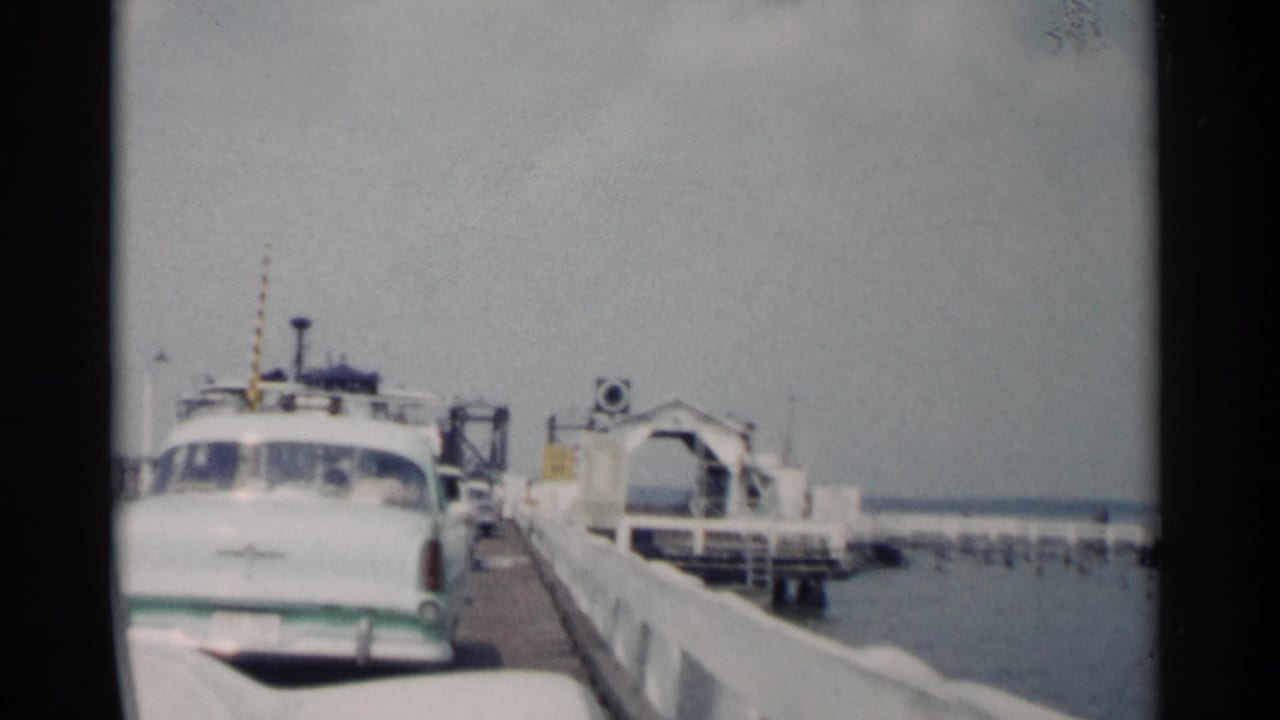 White mid-century sedan beside a ferry ramp at a waterfront dock.