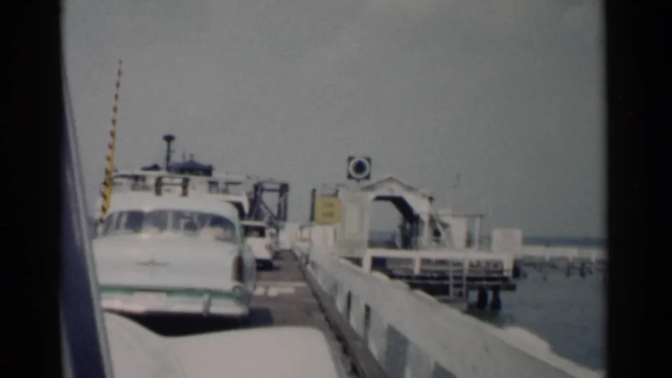 The home-movie frame shows a white mid-century sedan aligned near a ferry ramp, with dock railings, water, and small distant figures in the background.